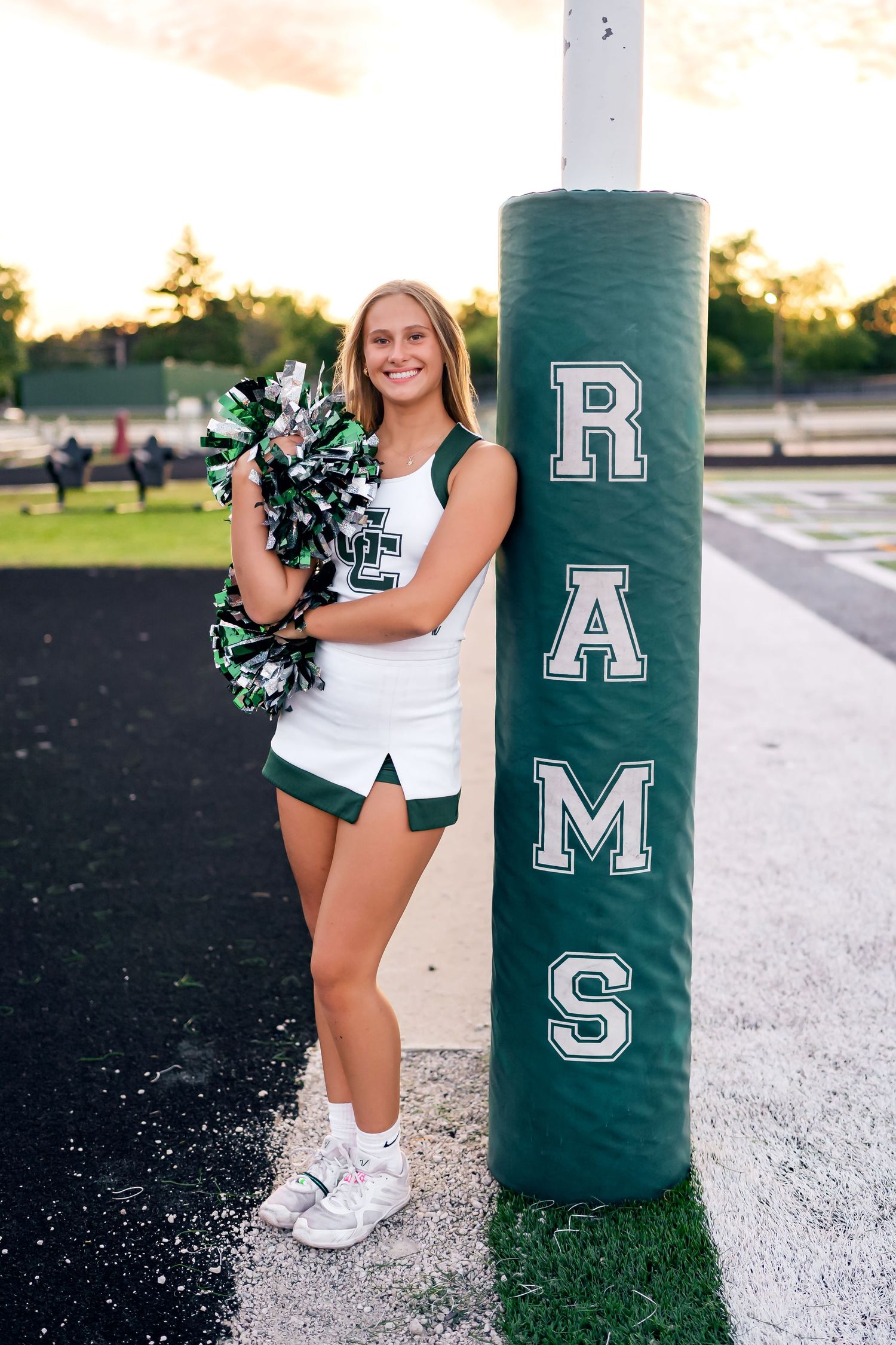Cheerleader in white uniform poses with green and white pom poms next to school mascot pole.