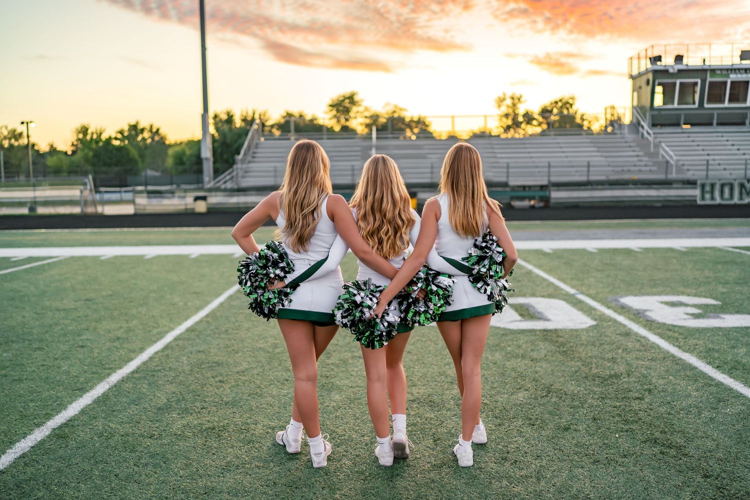 Three cheerleaders in green uniforms pose from behind on a football field at sunset.