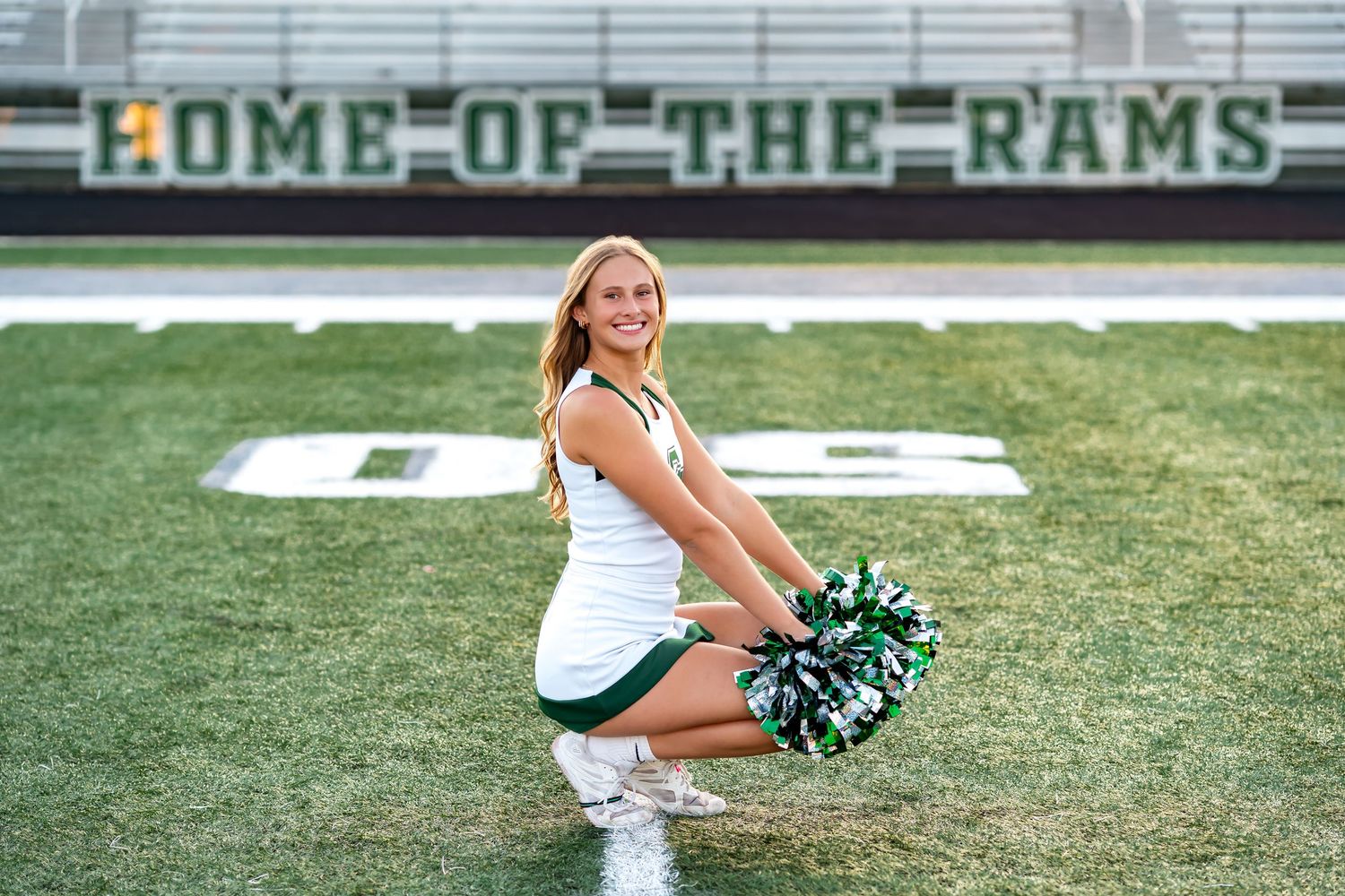 High school cheerleaders strike poses on a football field with the Home of the Rams scoreboard in background.