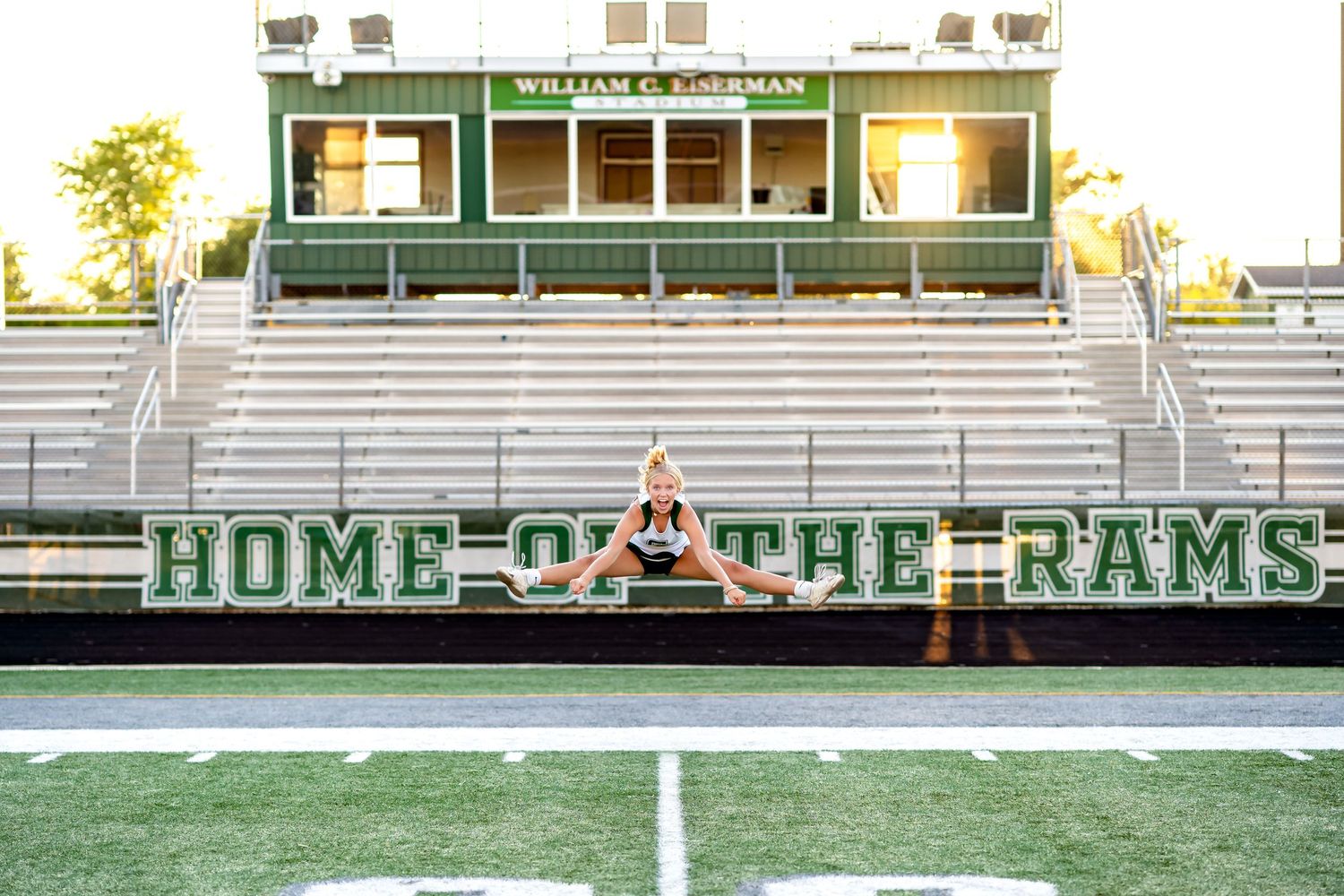 Football field with green artificial turf and HOME OF THE RAMS signage visible in white stadium bleachers at sunset.