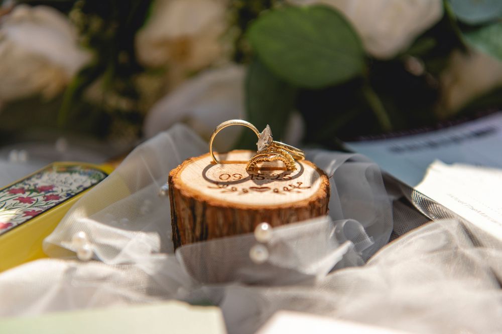 An elegant gold wedding ring with diamonds sits atop a light colored surface next to silk fabric and floral arrangements.