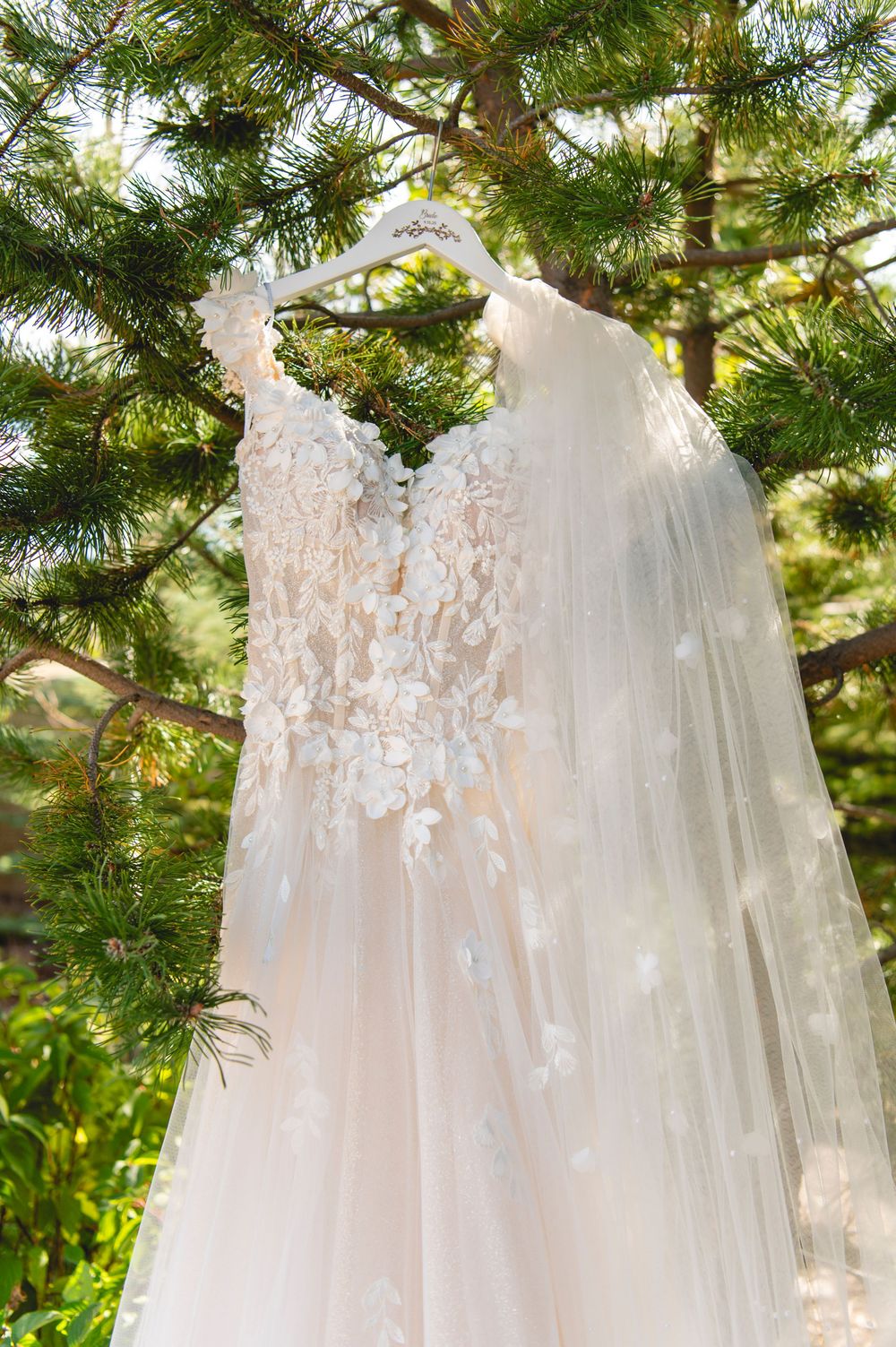 A close-up view of delicate lace and floral details on a white wedding dress hanging from evergreen tree branches.