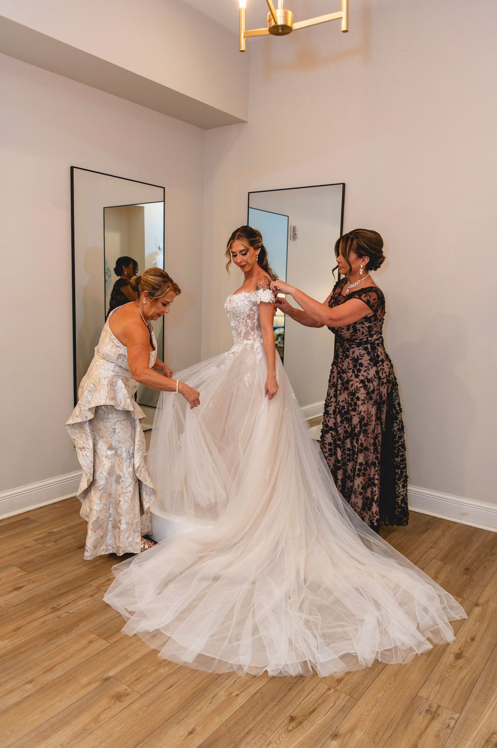 People preparing a wedding dress before a ceremony in a room with hardwood floors and off-white walls.