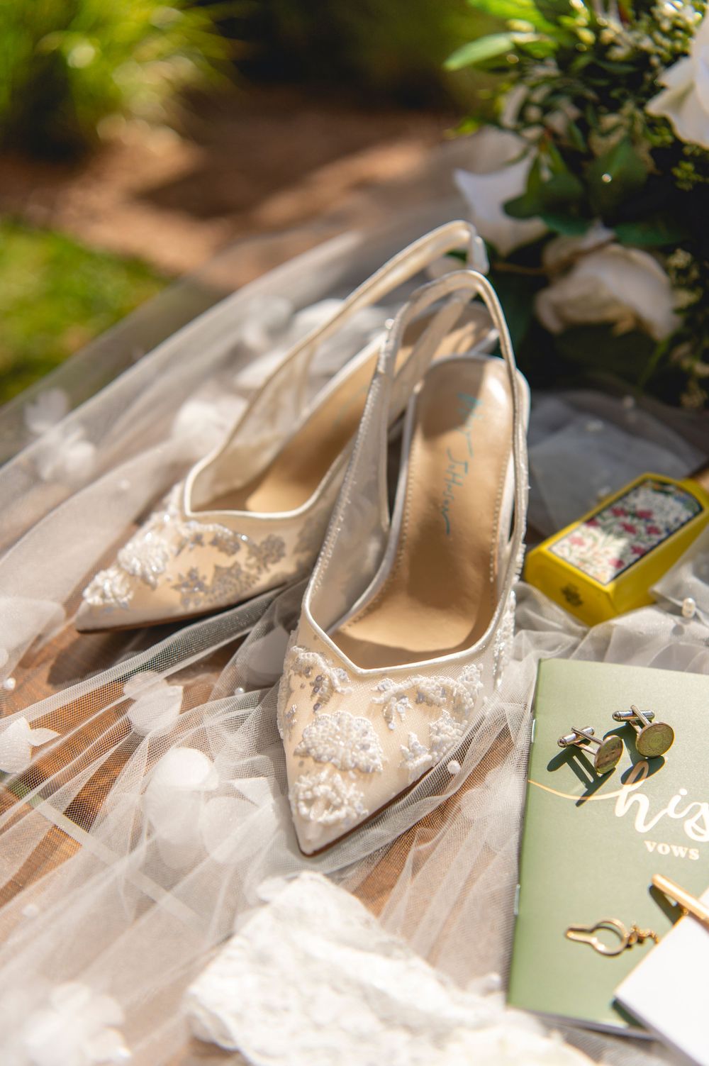 Elegant white lace wedding shoes displayed with a mint green wine bottle in dappled sunlight.