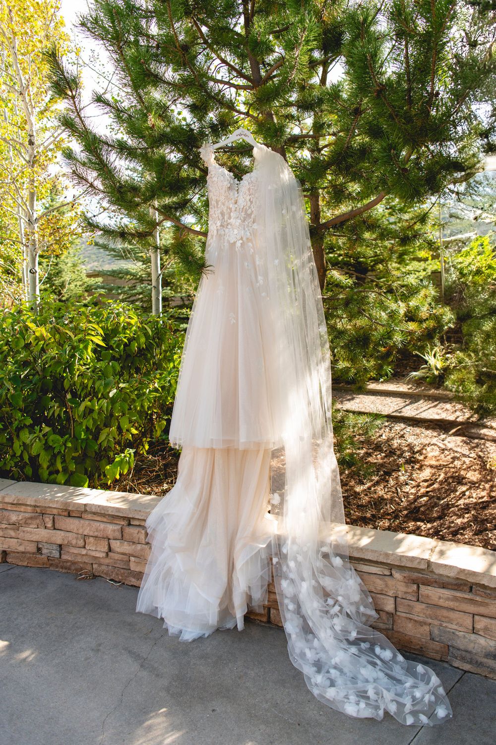 A white wedding dress with flowing layers of tulle hangs elegantly against a natural stone retaining wall in a garden.