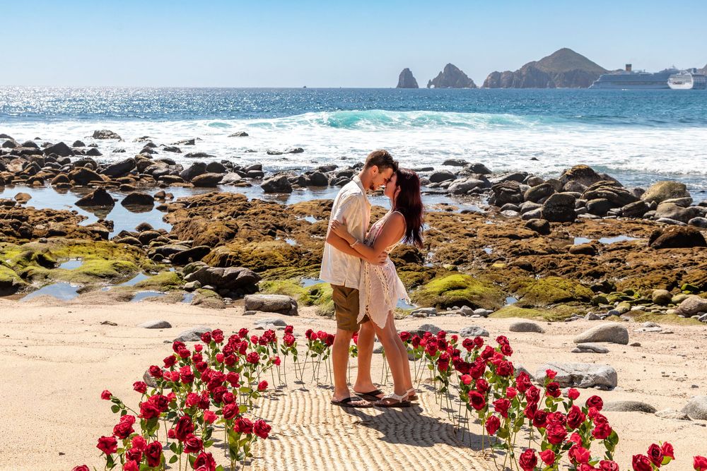 A heart shape made of red roses frames a romantic beach proposal scene at a scenic coastal location.