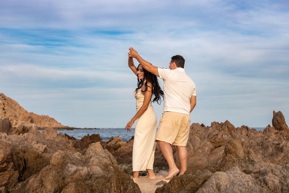 A couple sits together on coastal rocks overlooking the ocean during sunset in a romantic seaside setting.