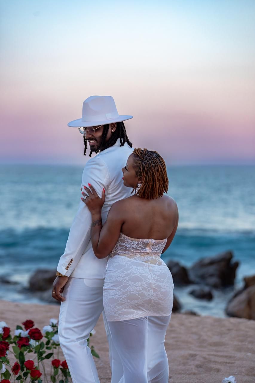 Couple in white wedding attire embracing on a sandy beach at sunset with red roses and ocean waves.