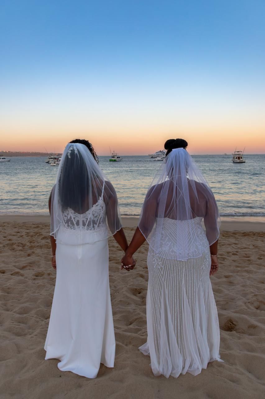 Two brides in white gowns and veils hold hands on a sandy beach at sunset with boats on the horizon.
