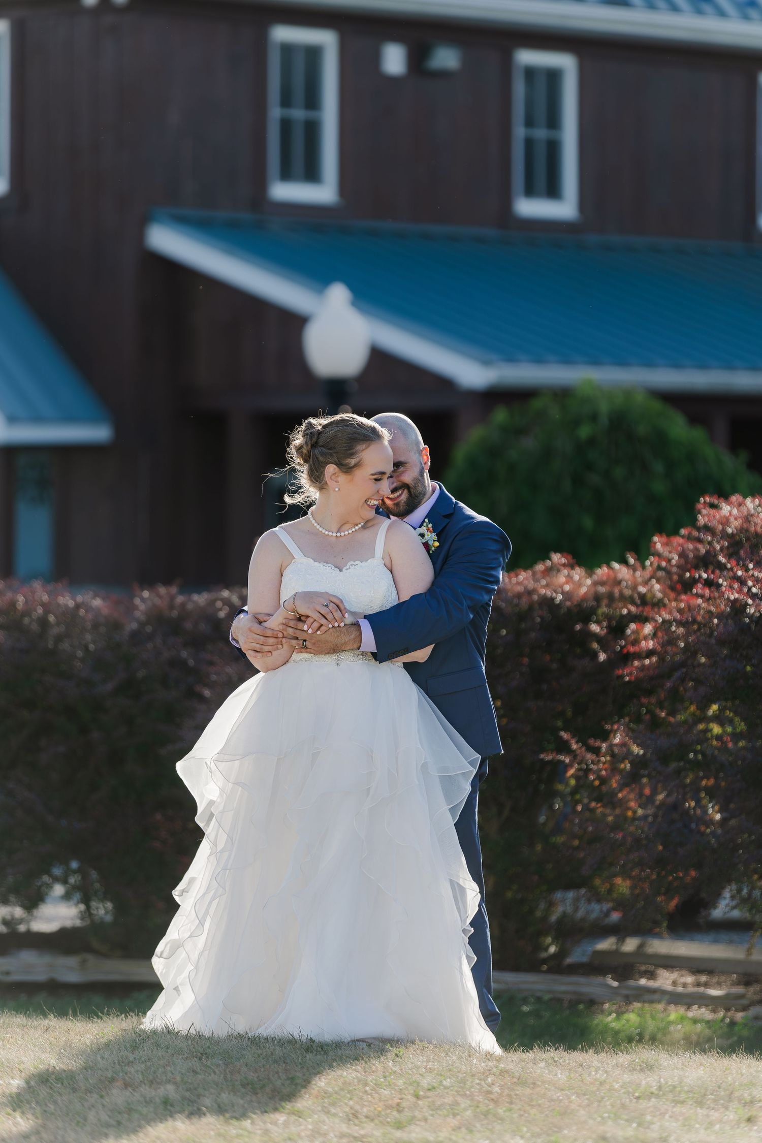 Groom embraces his bride in a tight hug from behind with the rustic building of Hauser Hall in Kitchener, Ontario behind them.