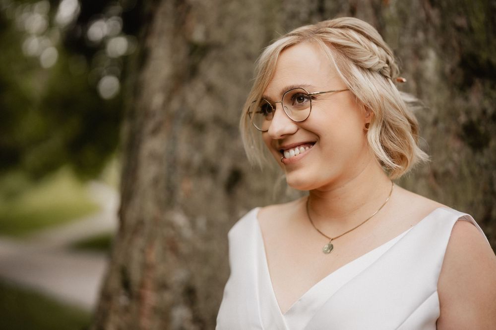 A portrait of a smiling person in a white dress wearing glasses and delicate jewelry.