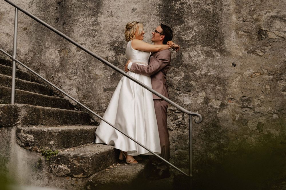 A couple shares an intimate kiss on rustic stone steps against a weathered wall while dressed in wedding attire.