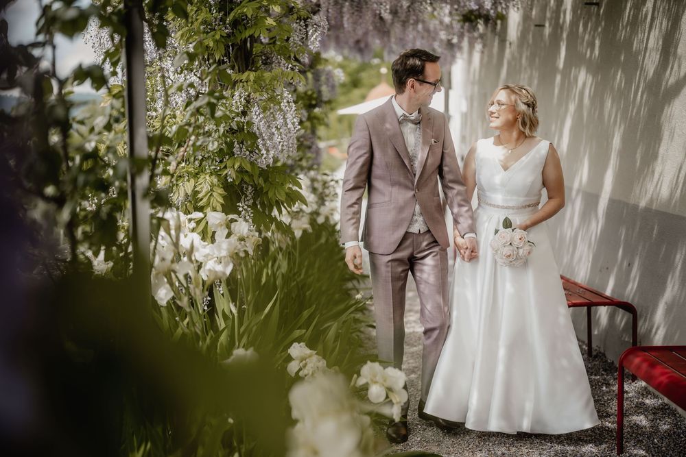 A romantic wedding photo of a couple in formal attire standing against a light wall surrounded by greenery.