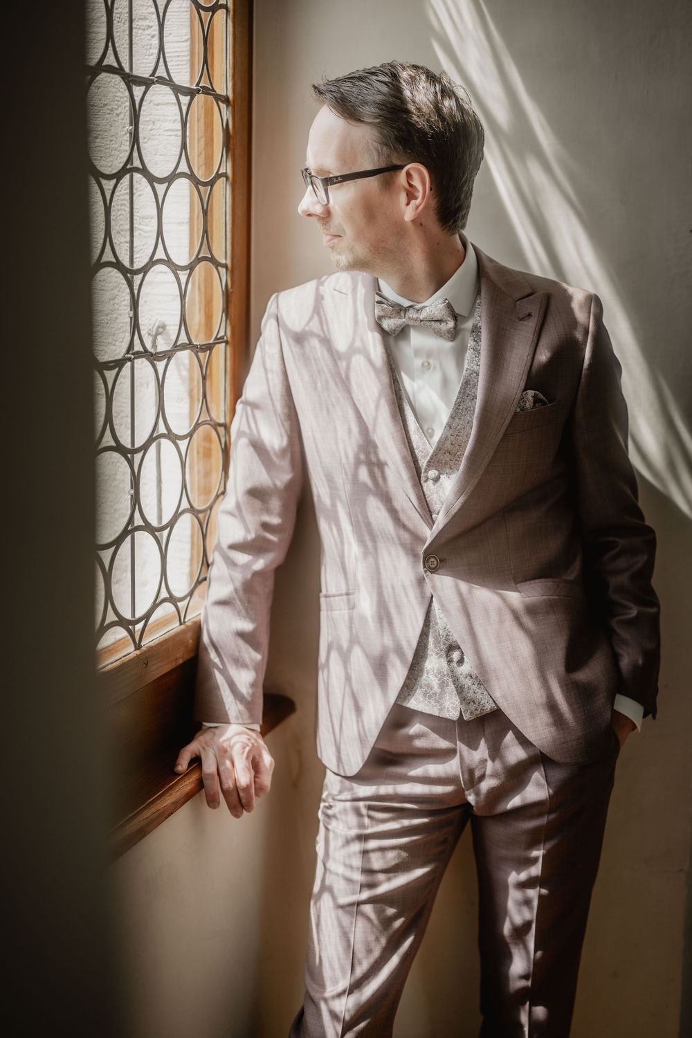 A person in a white suit gazes reflectively through a vintage window with ornate metalwork pattern.