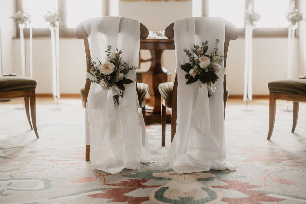 Two elegant white-draped chairs adorned with floral bouquets in a wedding ceremony setting.