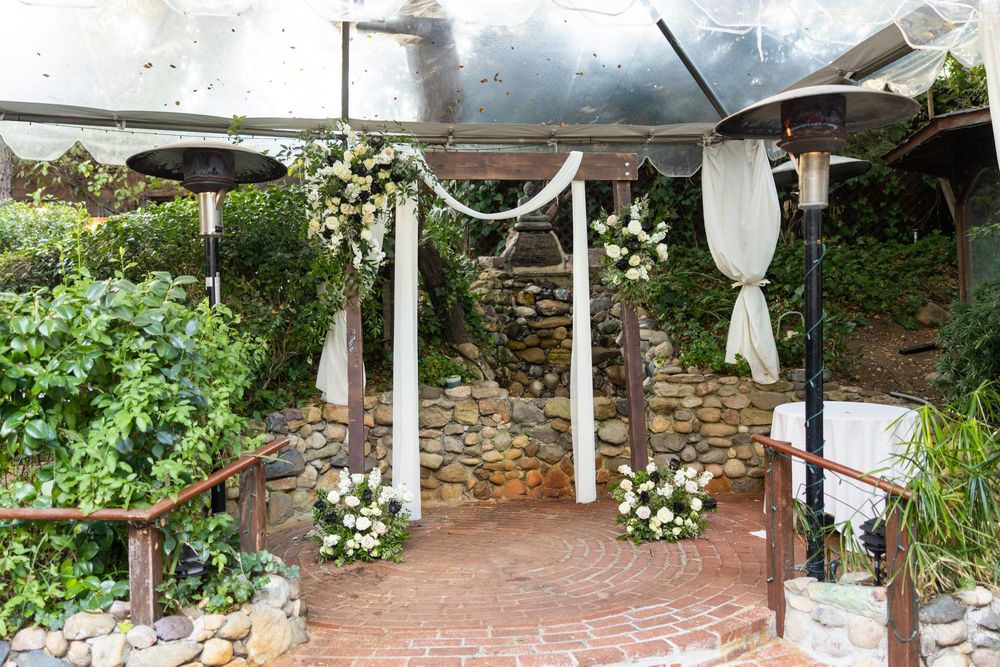 A panoramic view of a rustic outdoor garden path with stone walls, hanging white lanterns, and lush greenery.