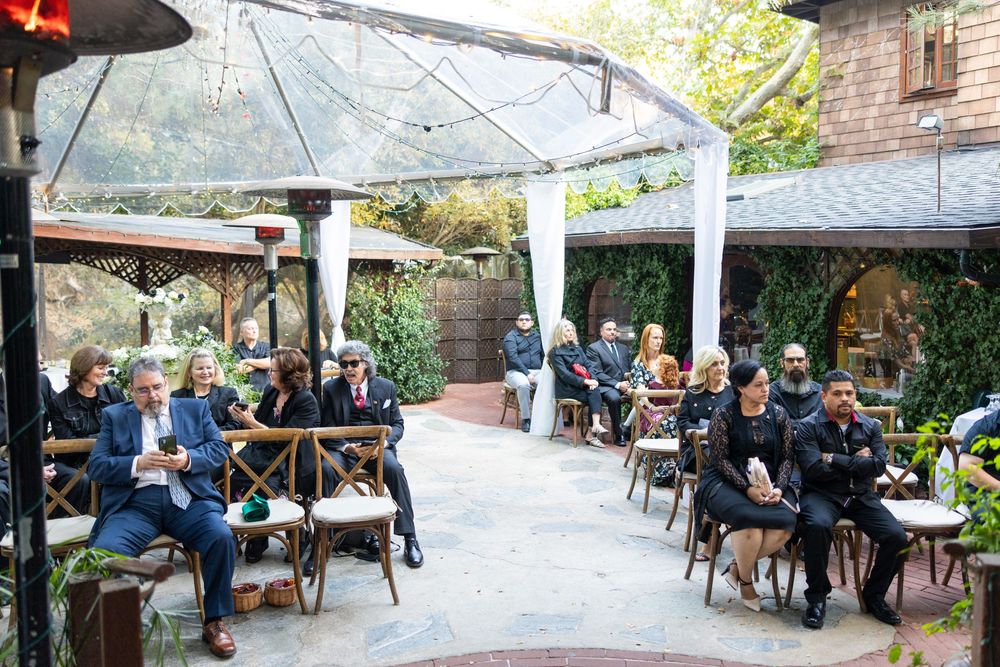 Diners enjoy outdoor seating under a covered patio space at a restaurant with brick walls and greenery.