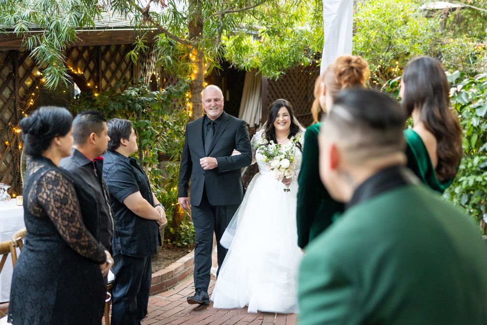 A group of wedding guests in formal attire gather in a lush garden setting during an outdoor celebration.