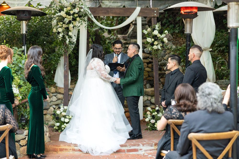 An outdoor wedding ceremony setup with wooden chairs and emerald green accents under a rustic covered patio.