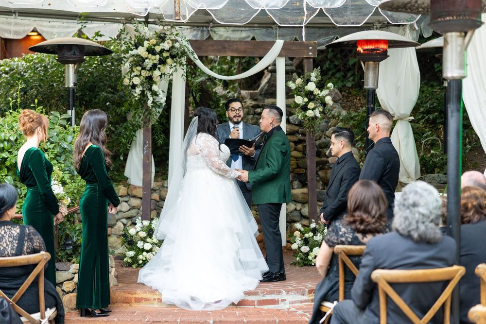 An outdoor wedding ceremony takes place under a decorated pergola with white drapes and floral arrangements.