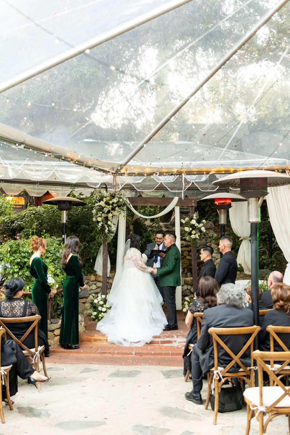 Wedding ceremony taking place under an elegant outdoor tent with wooden chairs and floral decorations.