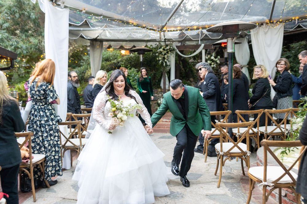 A couple joyfully exits their wedding ceremony at a rustic outdoor venue with string lights and wooden chairs.