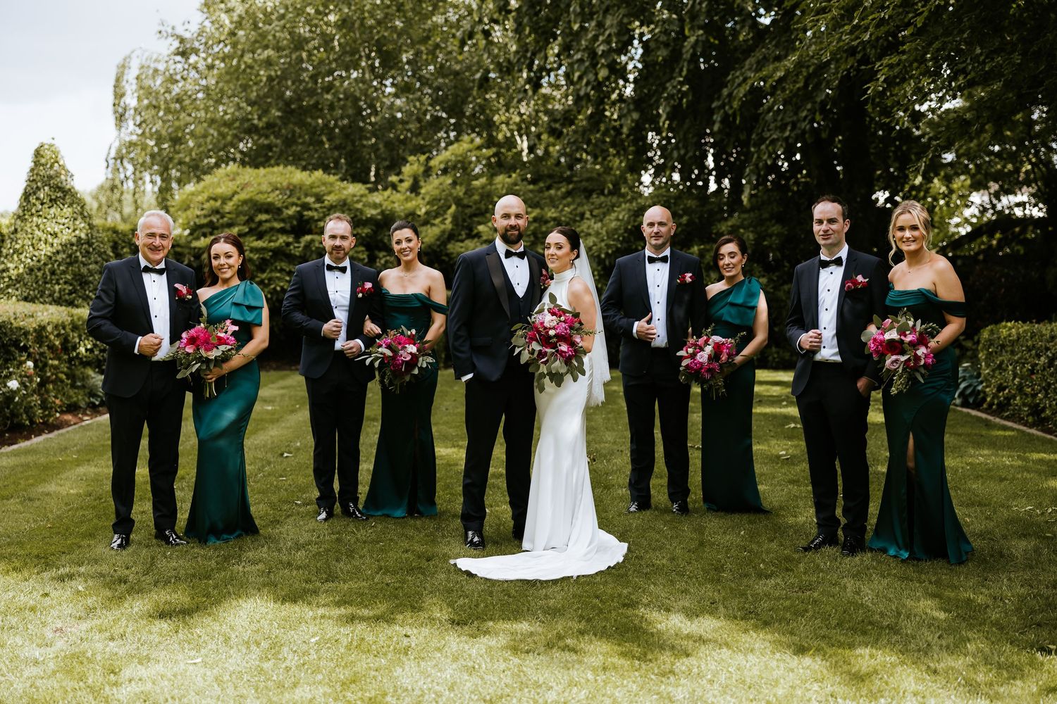 Wedding party in formal attire stands in a line on green grass during outdoor ceremony.