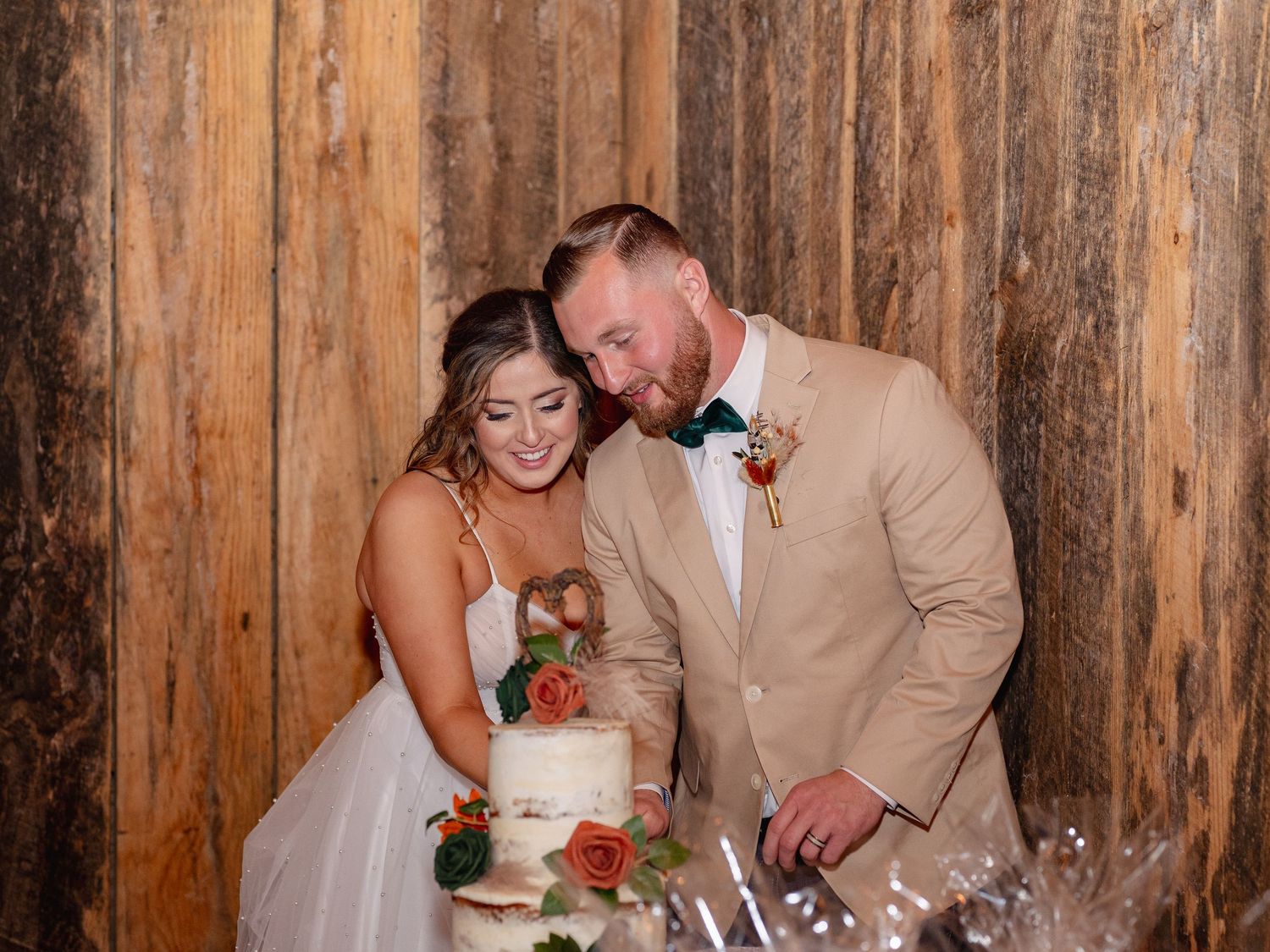 A newly married couple cuts their elegant white wedding cake with red roses at a rustic wooden-walled venue.