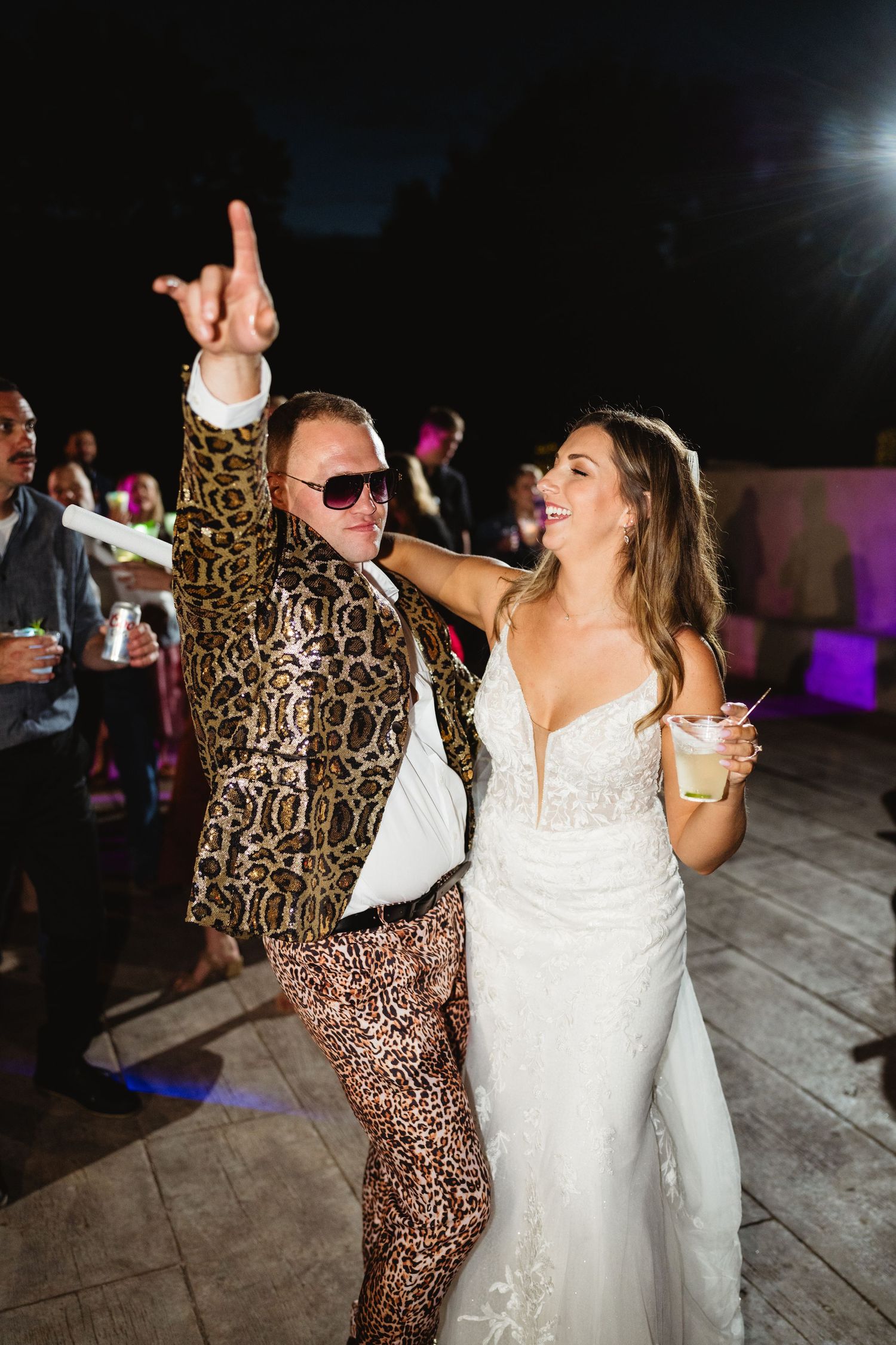 Wedding party guests dancing together on a wooden dance floor with colorful lighting in the background during reception.