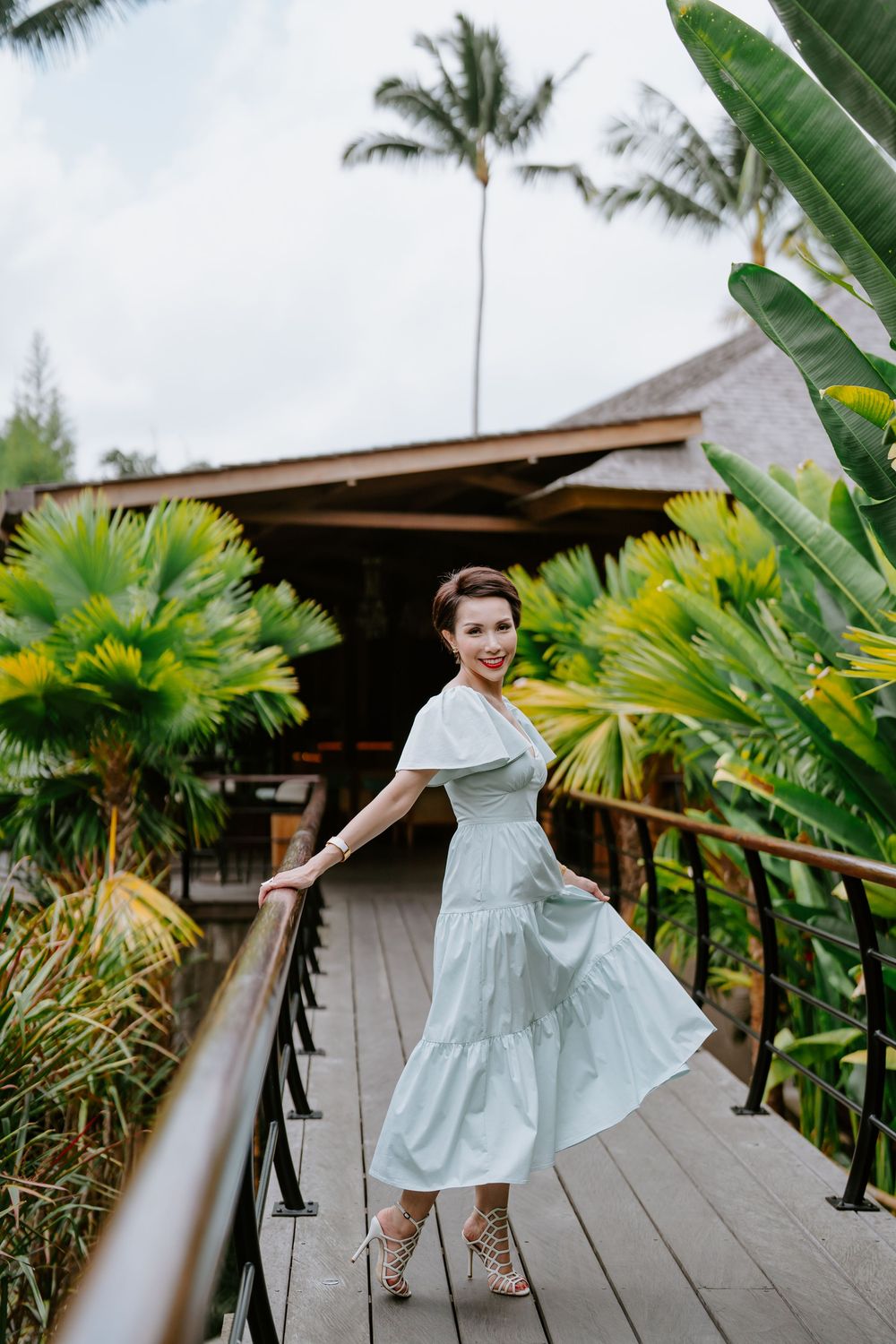 A light blue dress flows in the breeze on a wooden deck pathway with tropical landscaping.