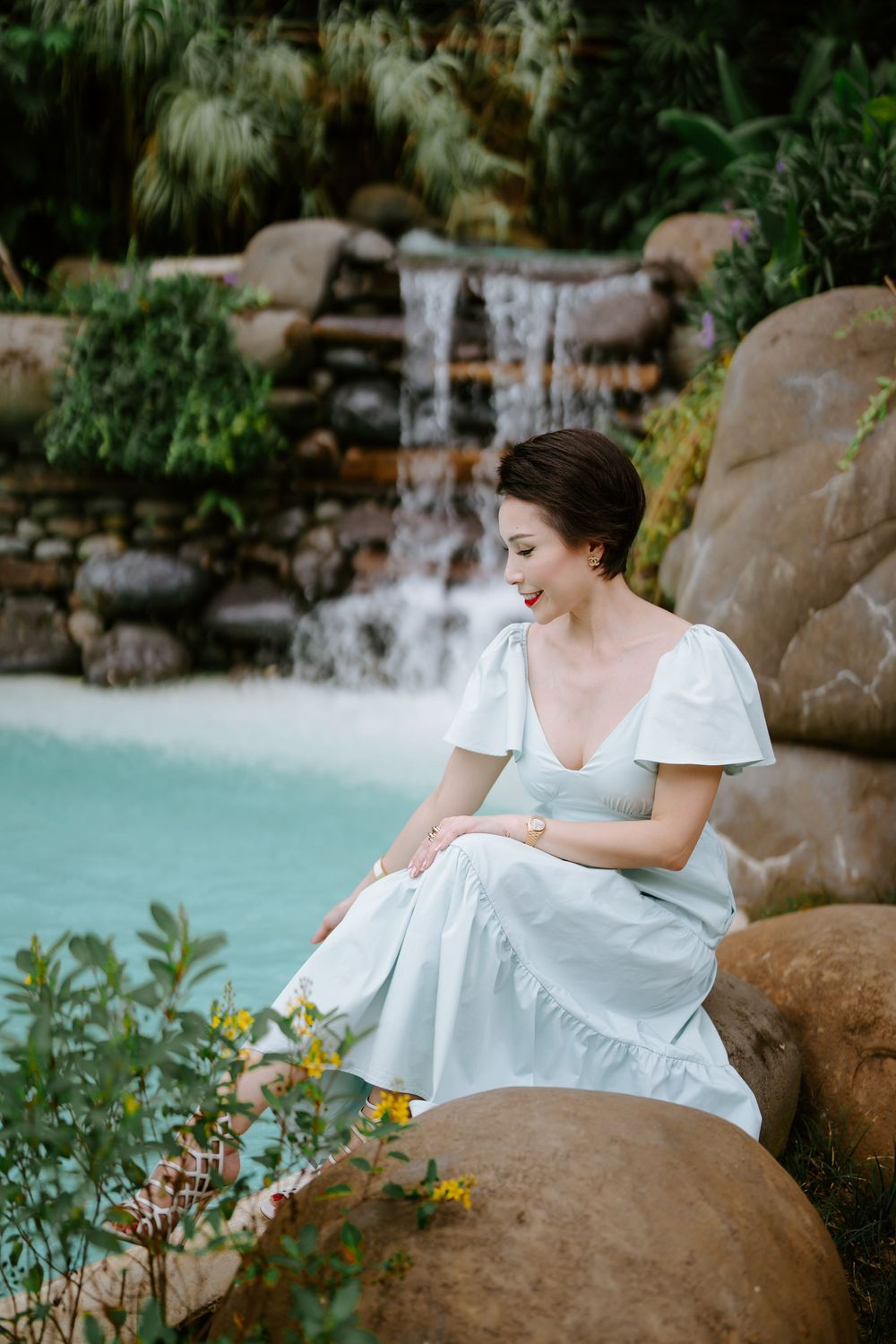 A serene poolside scene with a white dress against a rock waterfall and tropical flowers.