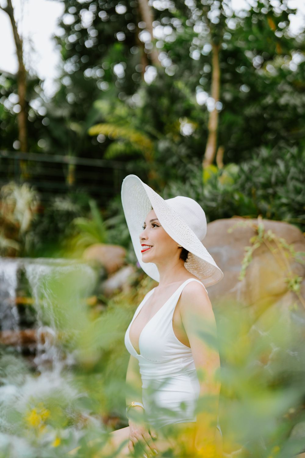 A white sunhat and swimsuit create an elegant silhouette among tropical foliage by a waterfall.