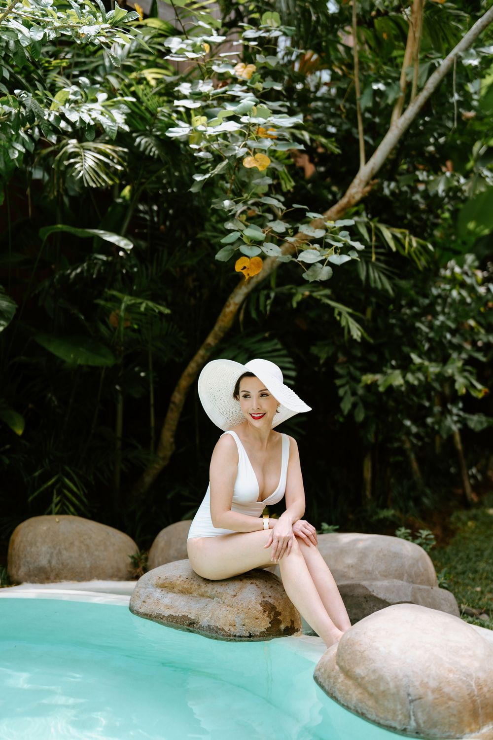 A classic white swimsuit and sunhat pose by a tropical pool surrounded by lush greenery.