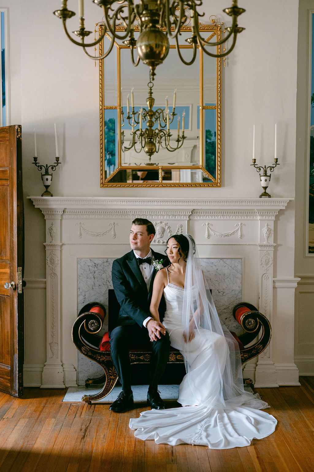 Elegant wedding couple seated on antique settee by ornate white fireplace with chandelier and mirror above.