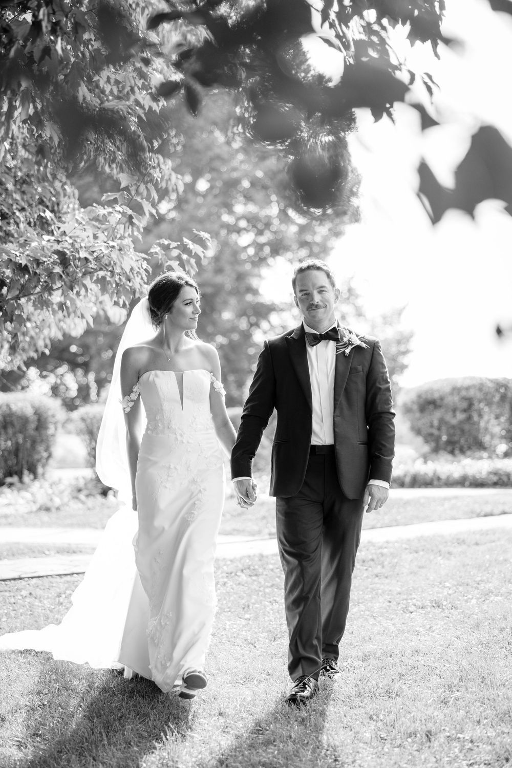 A romantic black and white wedding photo series showing a couple walking together through a sunlit garden.