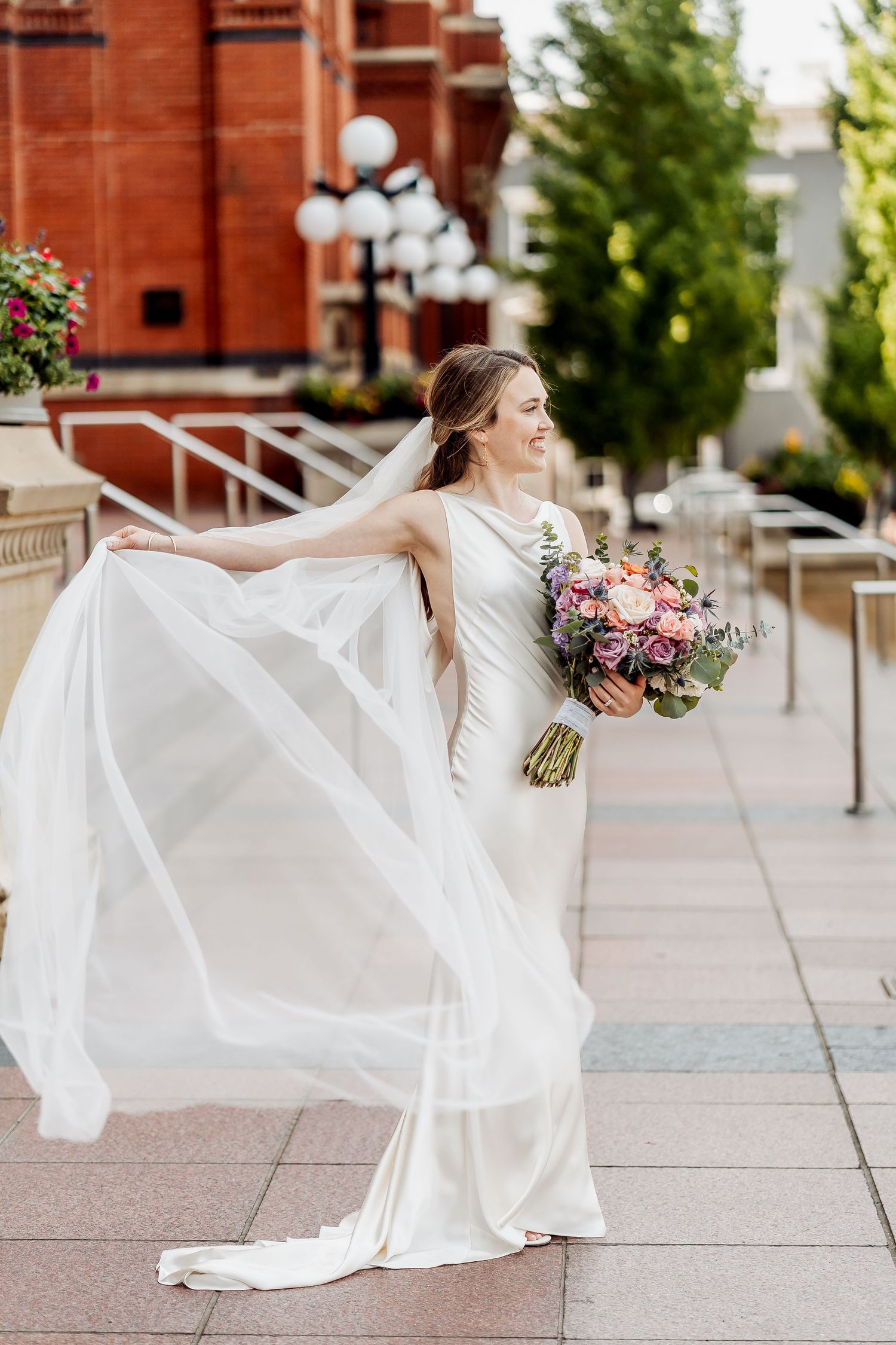 A bride in a flowing white dress spins gracefully on a plaza with red brick buildings in the background.