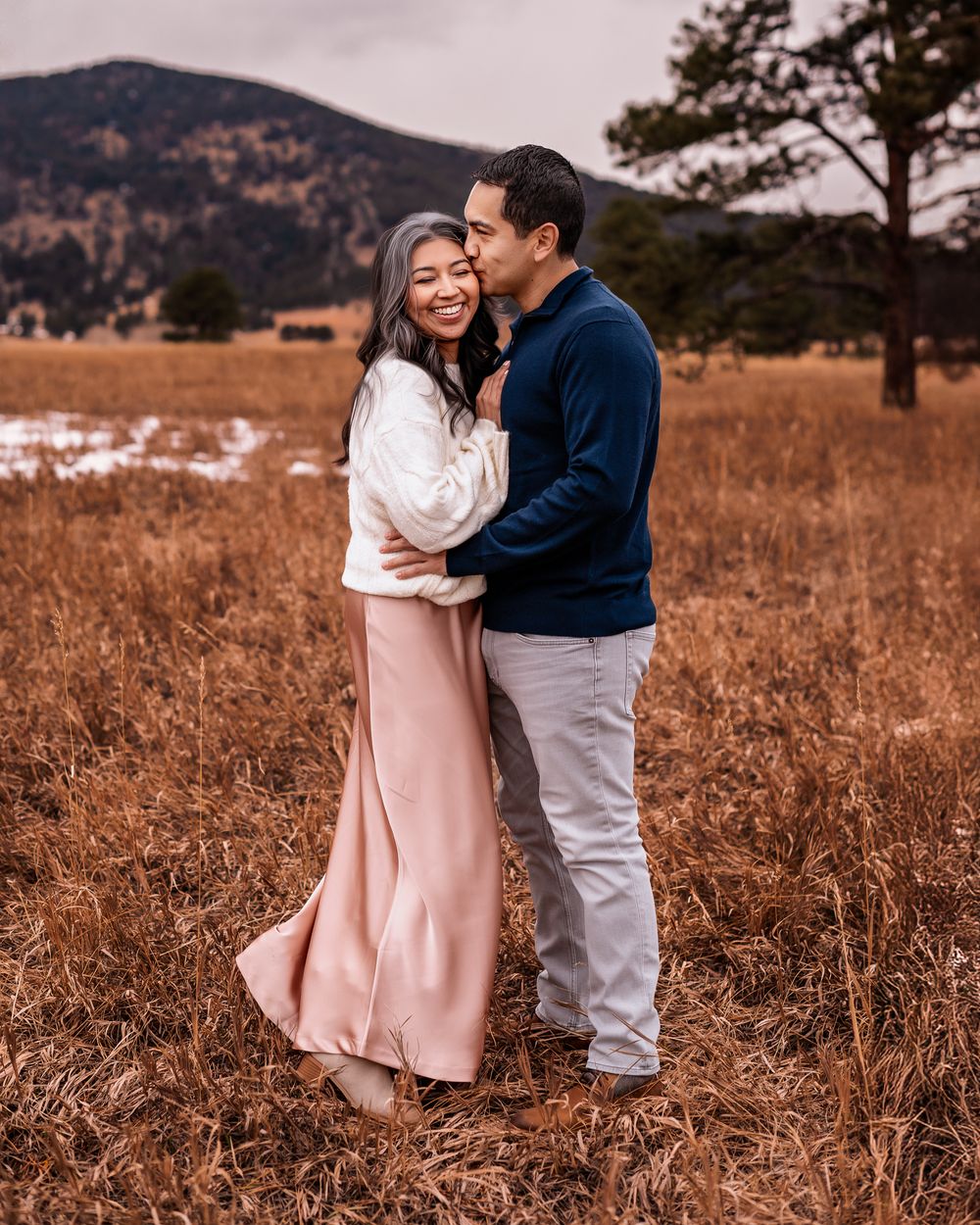 a husband kissing his giggling wife's cheek in front of an evergreen, CO mountain