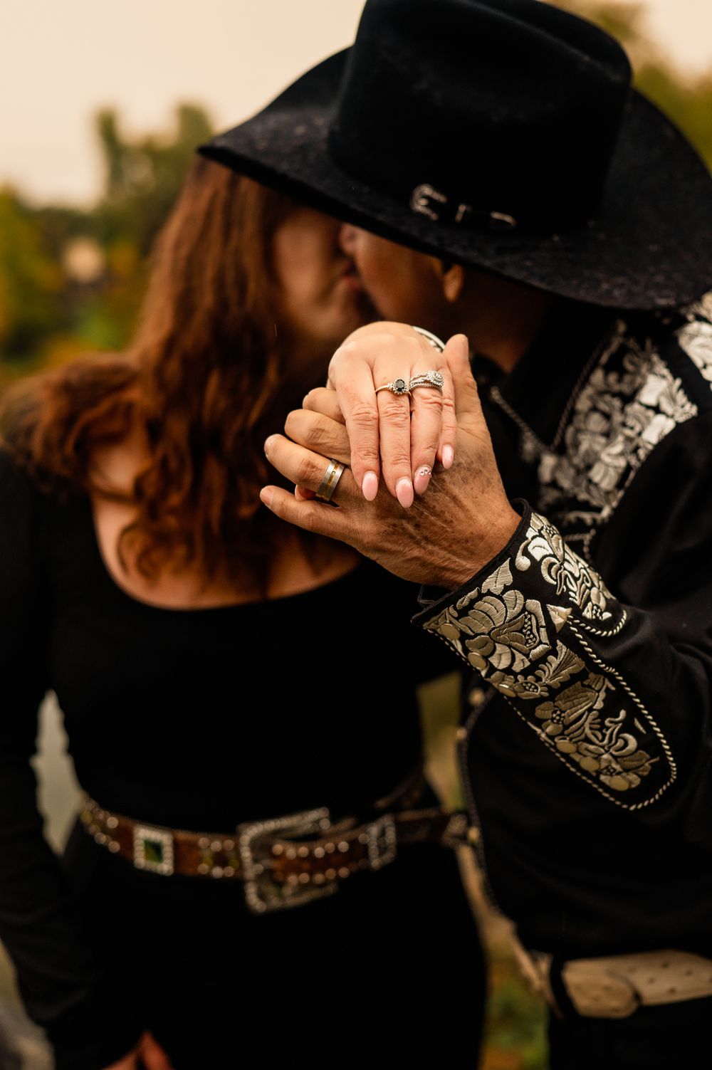 a husband holding his wife's hand up to the camera to show off her wedding ring while they kiss in cowboy attire