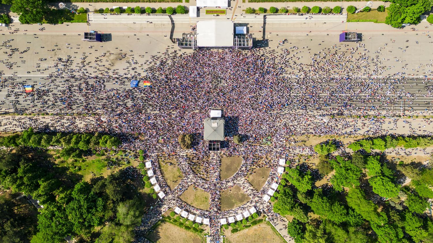 Aerial view of a massive outdoor concert crowd packed into a festival area surrounded by lush green trees.
