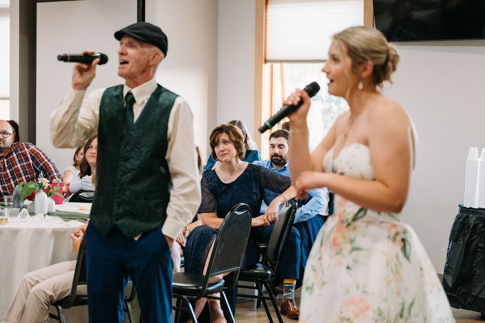 bride & dad singing karaoke, focus on mother of bride in background crying at Lowe Park in Marion Iowa taken by Midwest wedding photographer