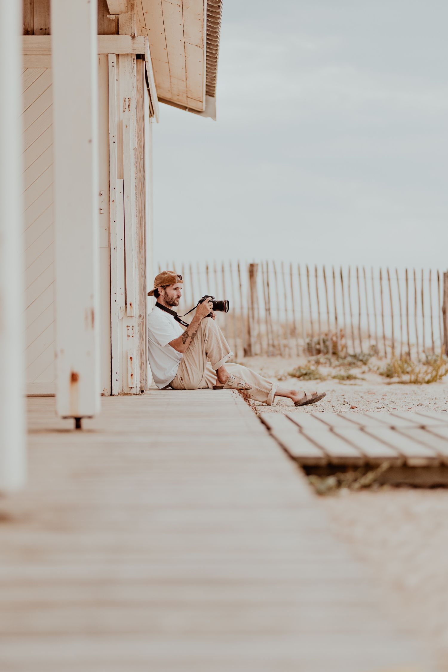 A photographer sits on a wooden deck near a beach fence capturing photos in warm sunlight.