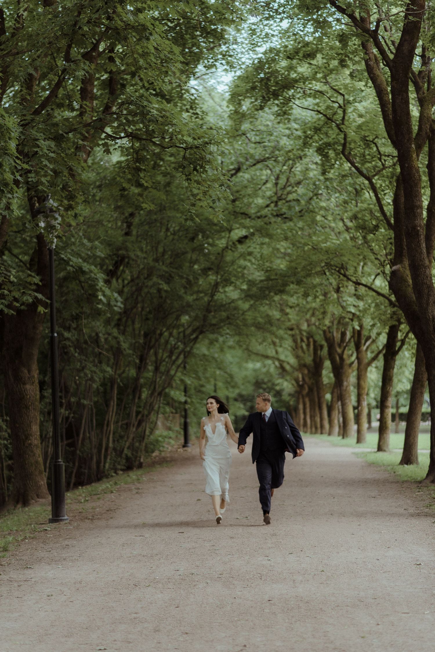 A couple running together on a tree-lined path in a lush green park setting.