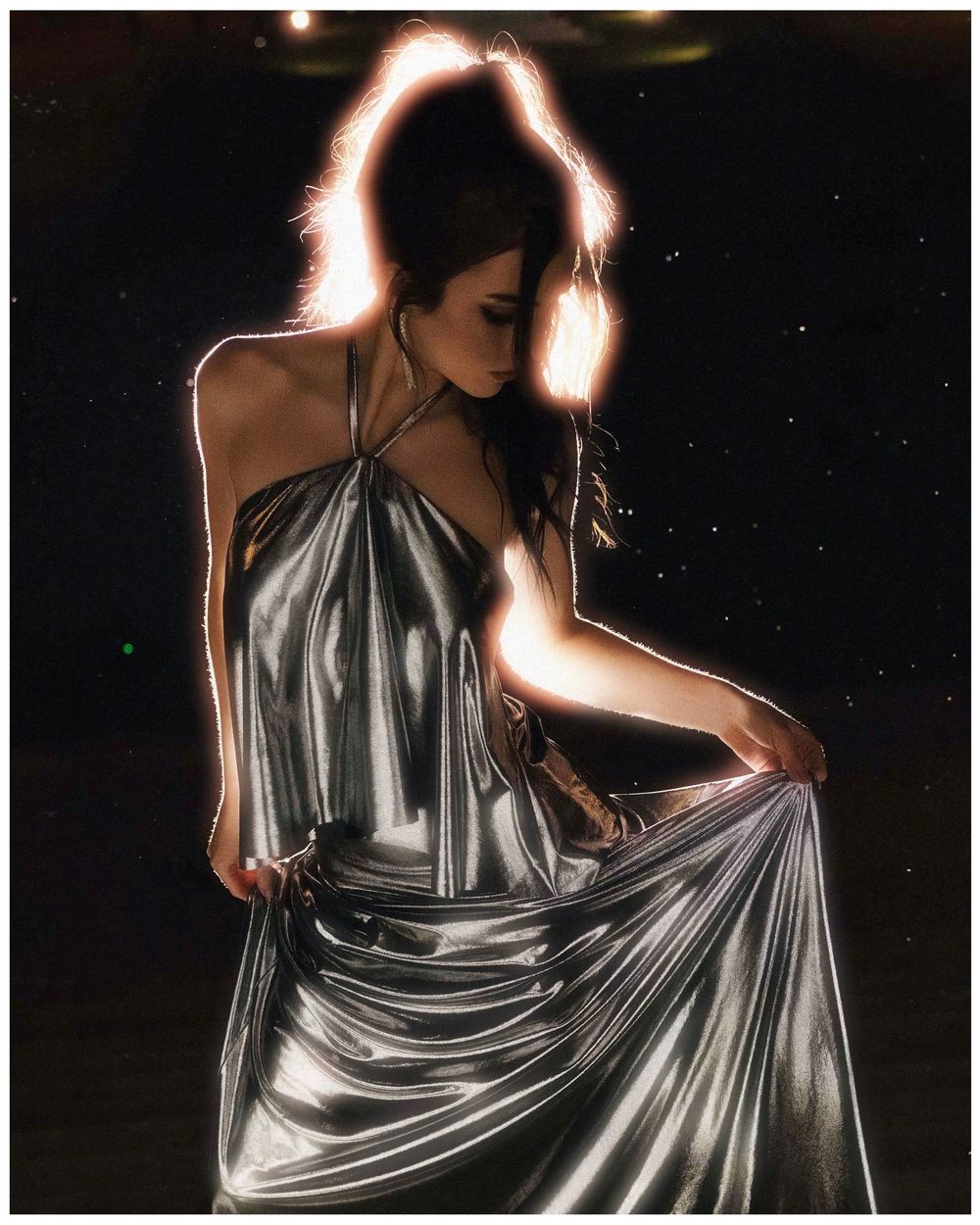 Woman in a metallic silver halter dress illuminated with backlit glow effect against dark background.