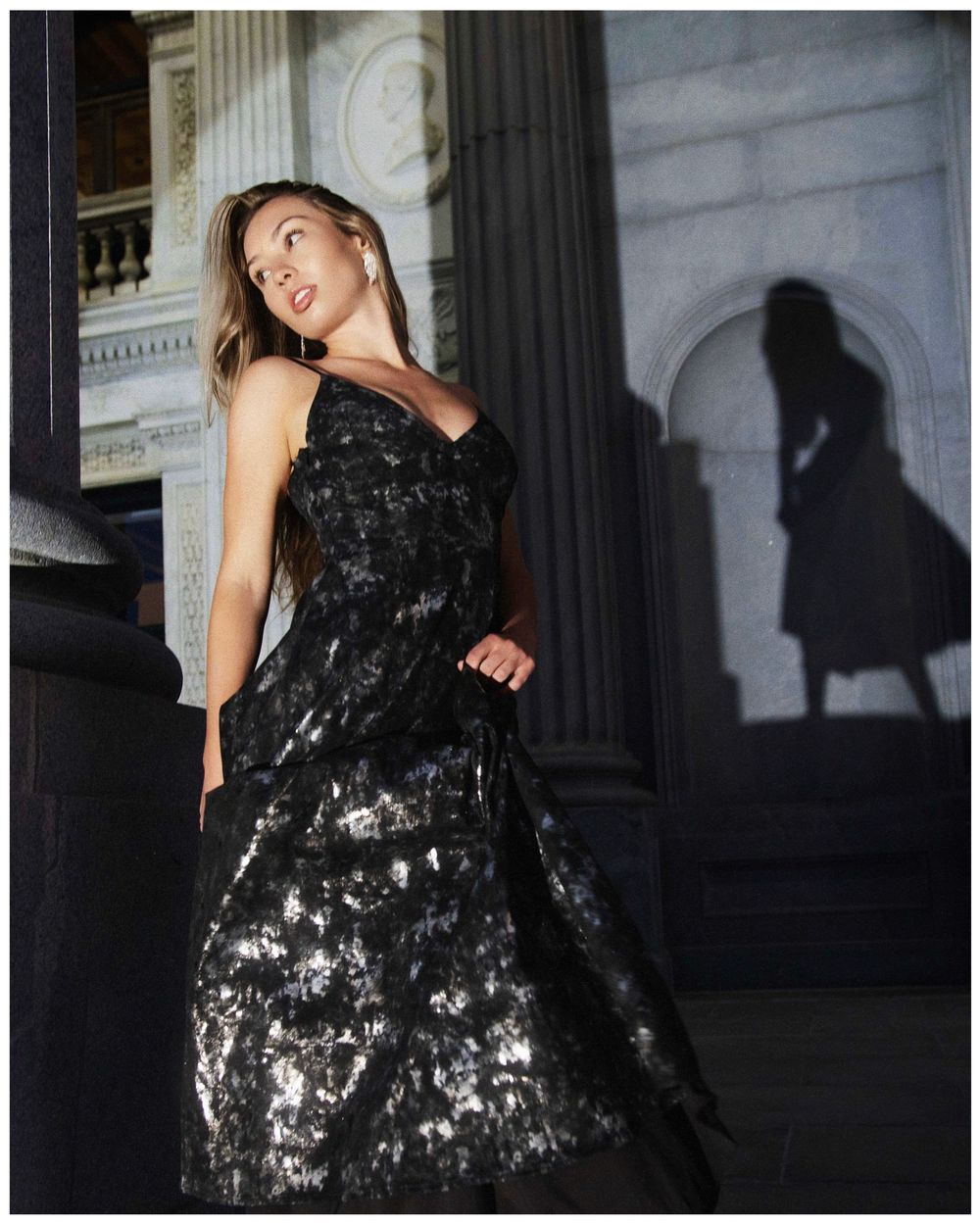 A sparkling black evening gown catches light while being modeled against architectural columns in moody lighting.