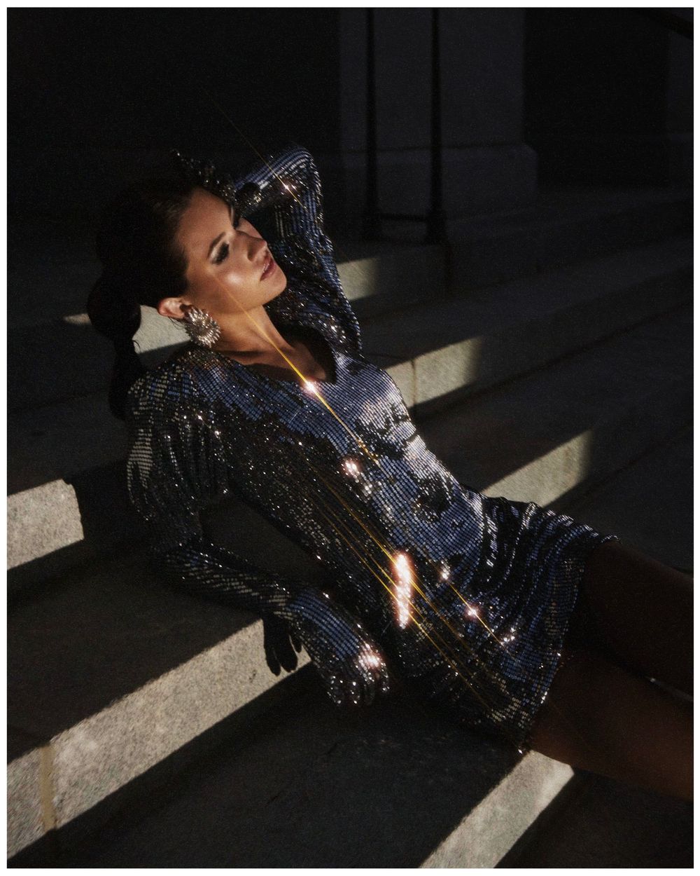 A person in a dark room wearing a sequined dress lying down as light streams in through a window, creating a dramatic mood.