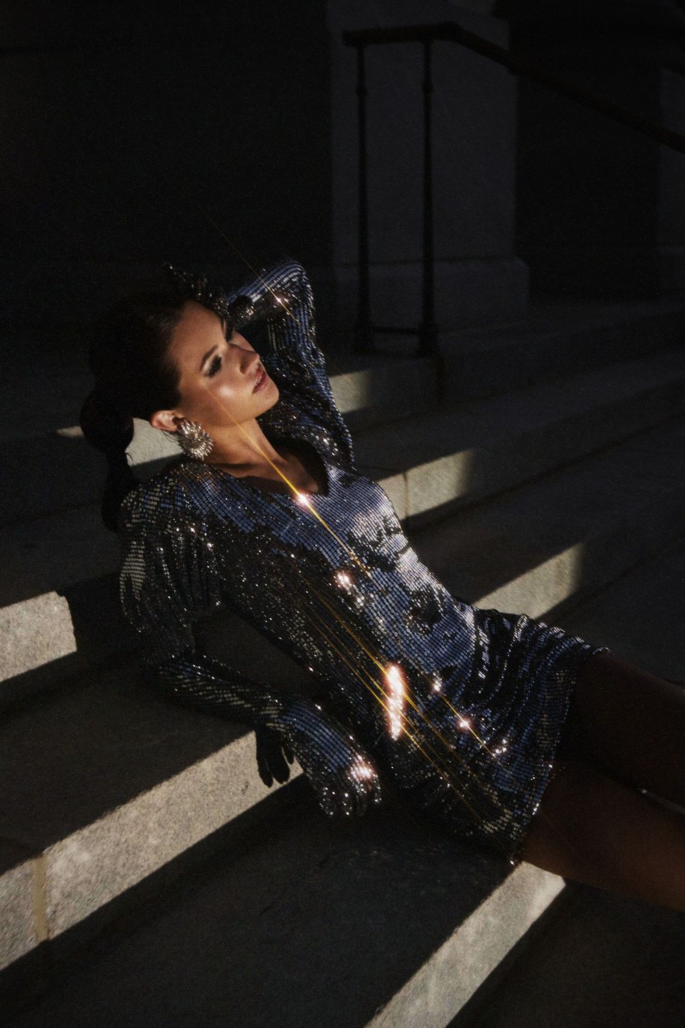 Fashion model in black sequin dress lies dramatically on floor creating artistic shadows in dramatic lighting.