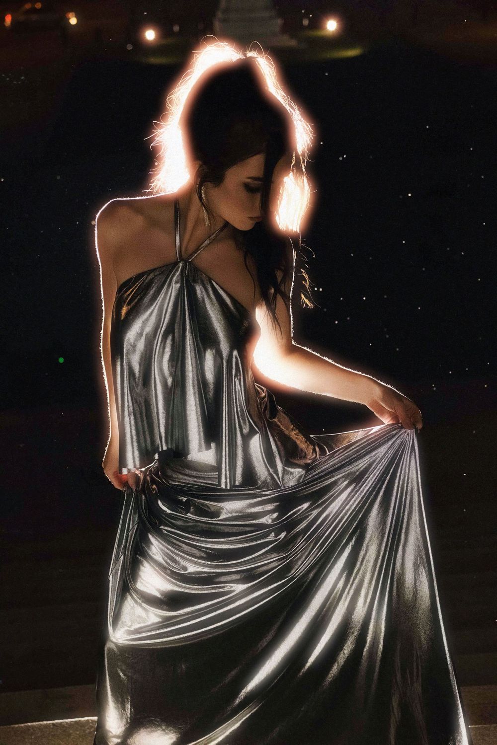 A dramatic series of backlit silhouettes show a figure in a shimmering silver halter dress twirling against a dark background.
