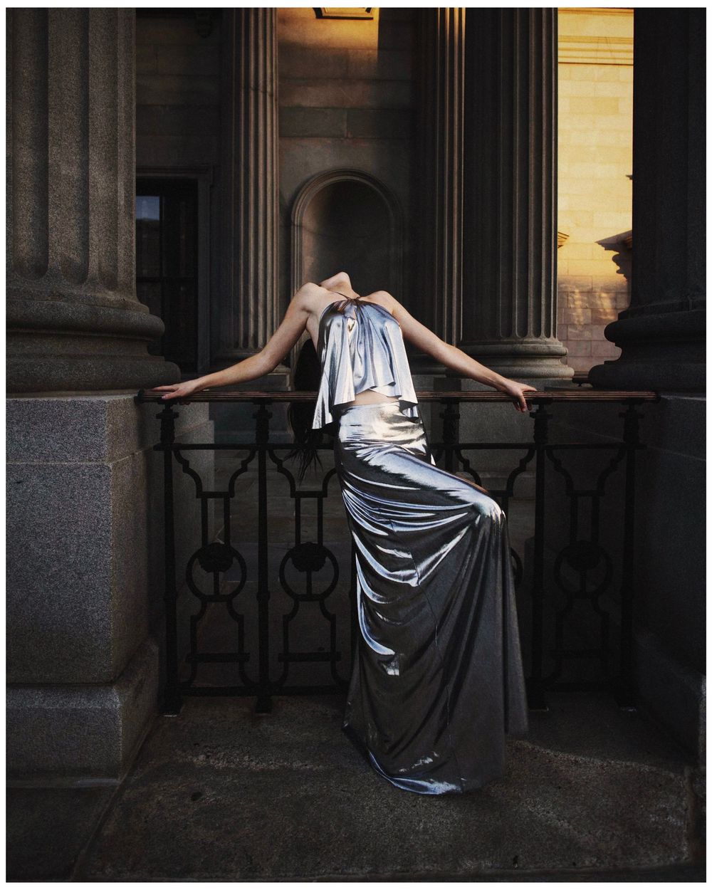A figure in a silver evening gown poses dramatically against stone columns at dusk.