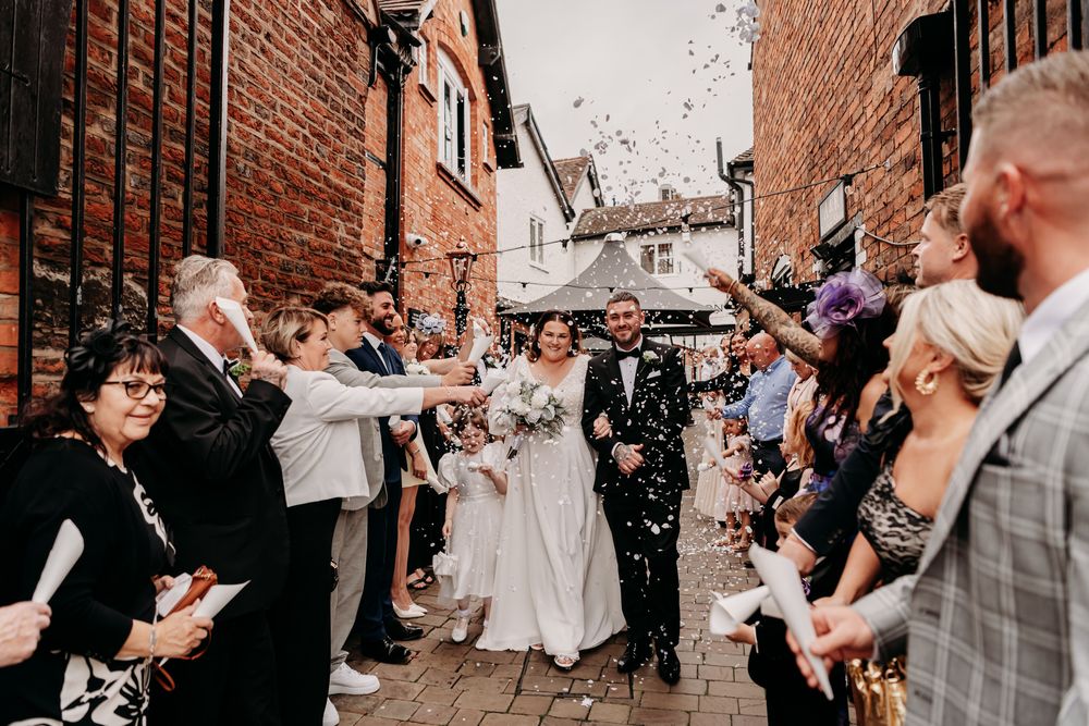 Wedding guests throw white confetti on a couple along a rustic brick wall during a celebration.