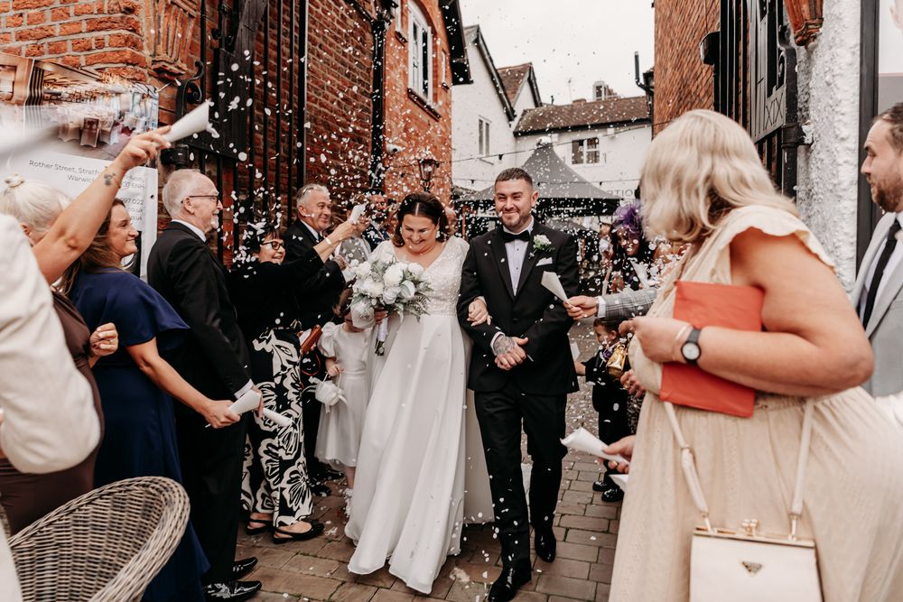Wedding guests stand along charming brick walls in a historic street celebration.